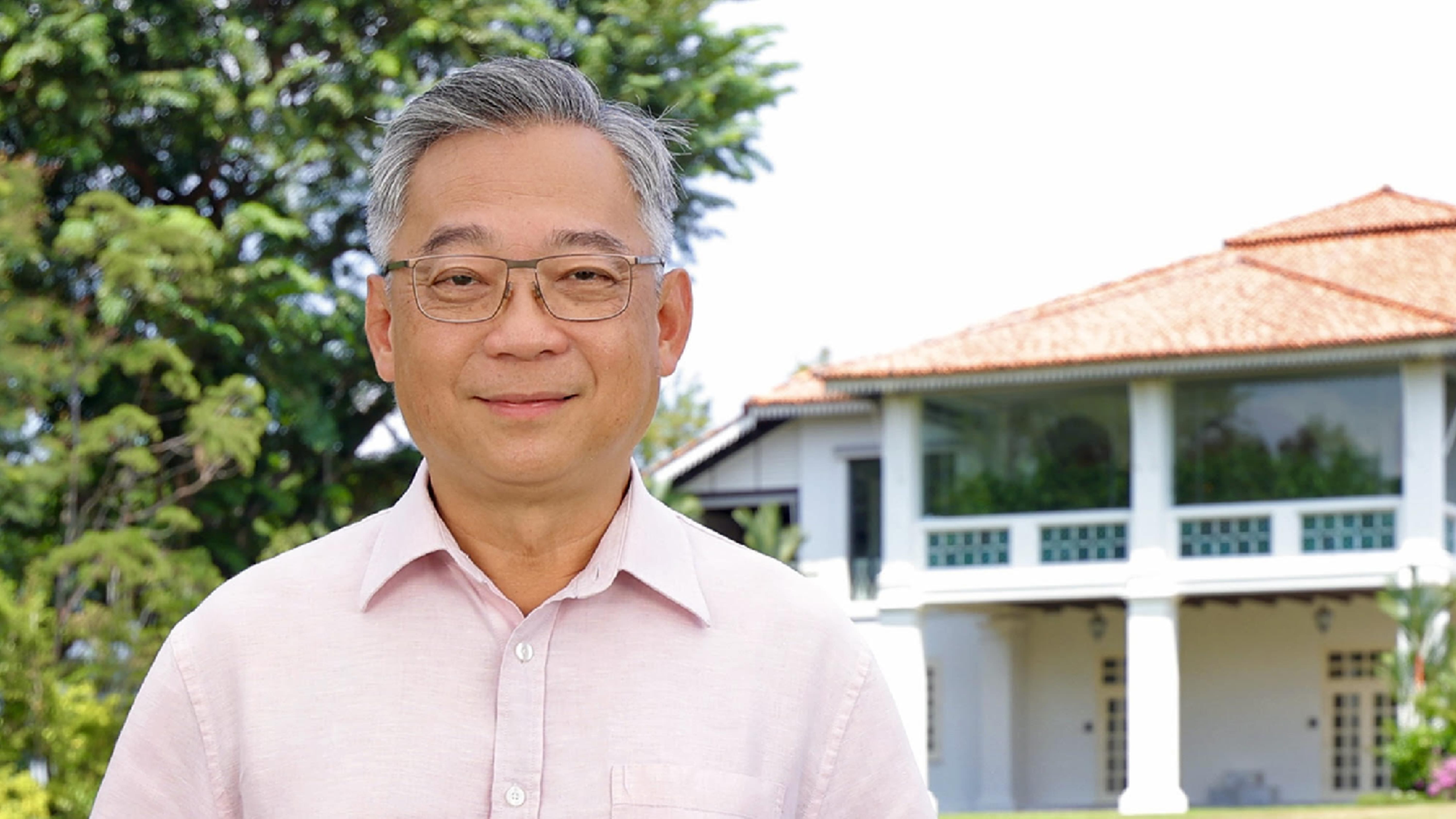 Man in pink shirt and glasses, stands outside of a white building with an orange roof.